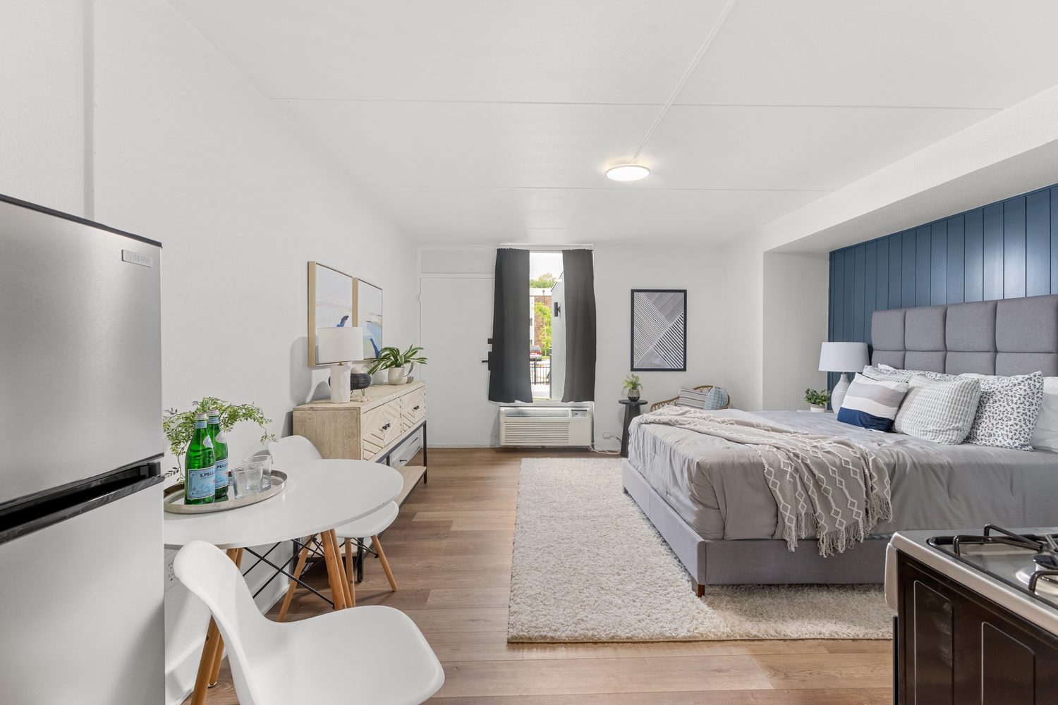 A staged studio apartment with a queen bed against a navy panelled wall, a round table for two, and wide-plank wood-look flooring.