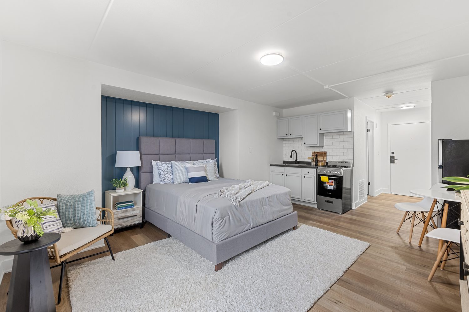 A staged studio apartment with a queen bed against a navy panelled wall, a round table for two, and wide-plank wood-look flooring.